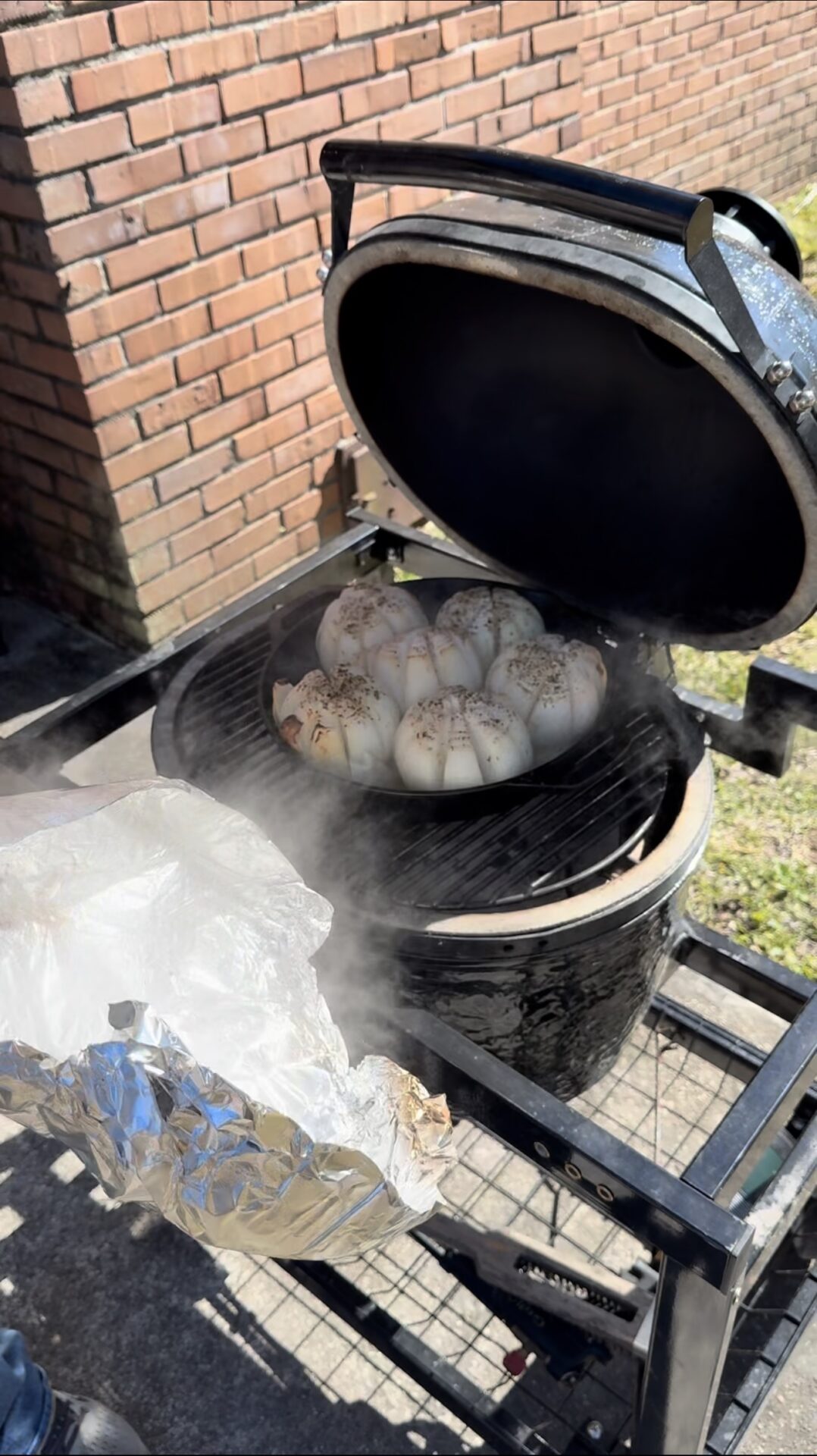 A cluster of whole garlic bulbs is being smoked on a grill with the lid open, releasing steam. A person’s hand is holding a piece of foil nearby. The grill is outside next to a brick wall.