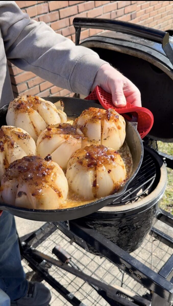 A person holds a cast iron skillet with six whole onions topped with melted cheese and seasonings, freshly cooked on an outdoor grill. The onions are golden and slightly caramelized.