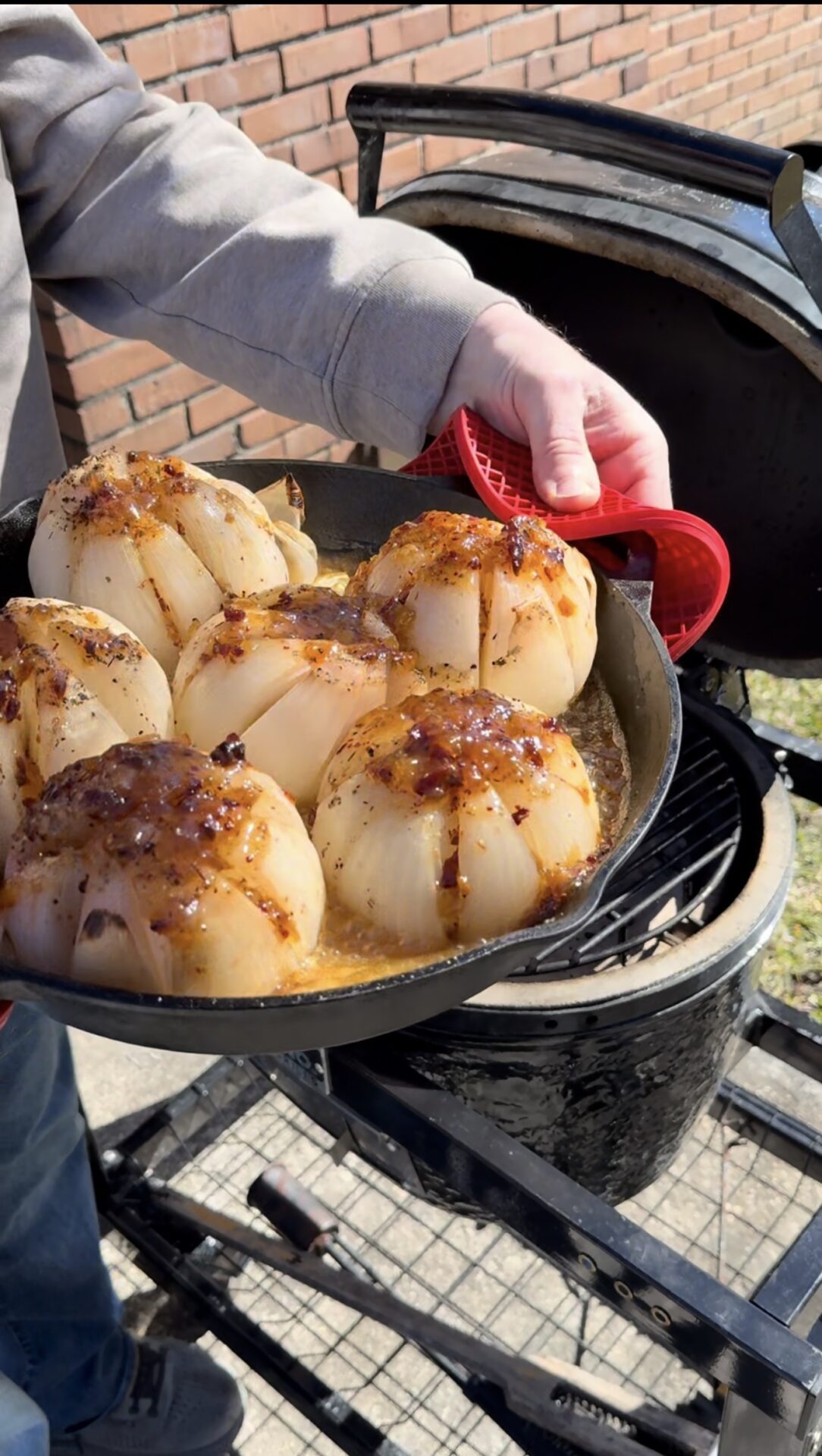 A person holds a cast iron skillet with six whole onions topped with melted cheese and seasonings, freshly cooked on an outdoor grill. The onions are golden and slightly caramelized.