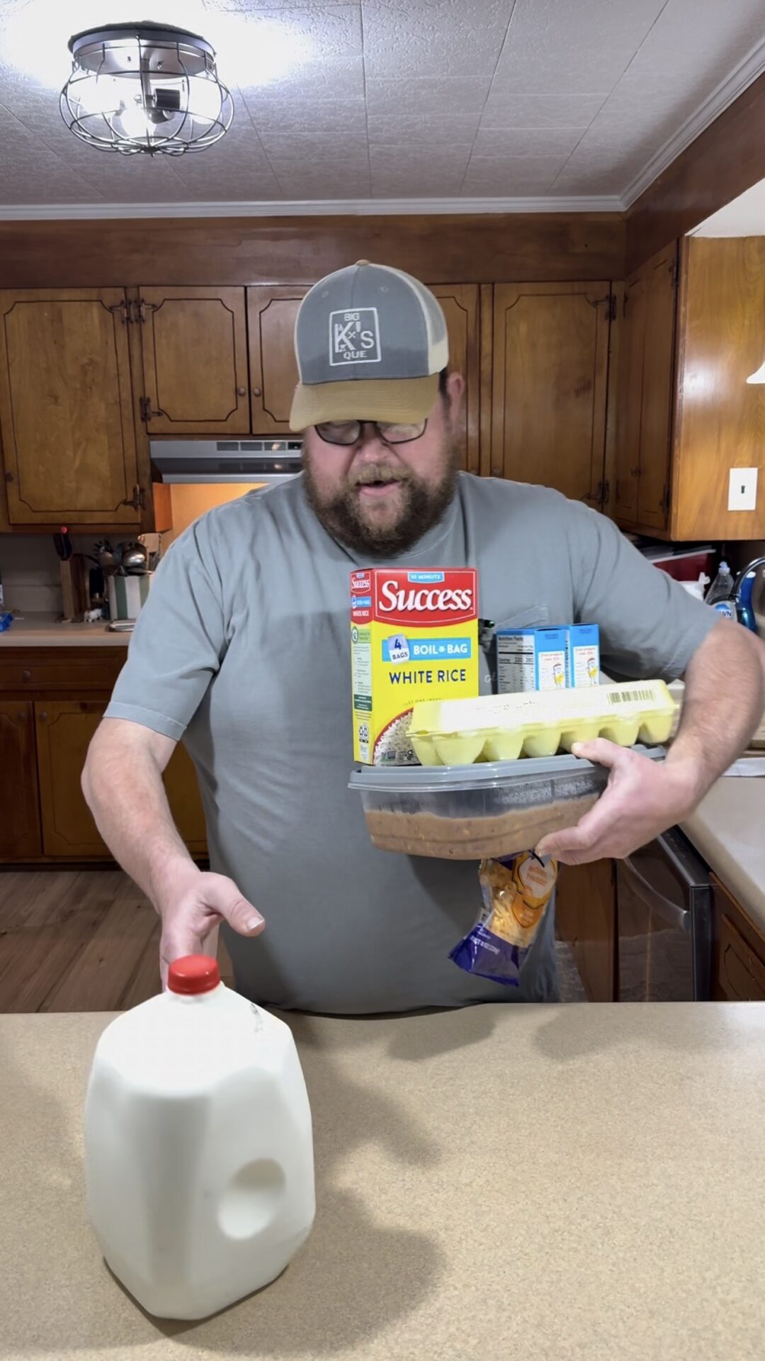 A bearded man in a gray shirt and cap balances groceries, including eggs, rice, and canned goods, in one arm while reaching for a jug of milk on a kitchen counter. The kitchen has wooden cabinets and a light ceiling.