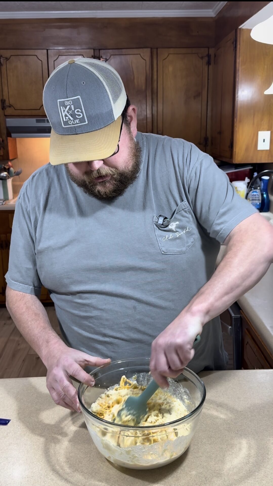 A man wearing glasses, a gray T-shirt, and a gray cap mixes ingredients in a glass bowl with a green spatula in a kitchen with wooden cabinets.