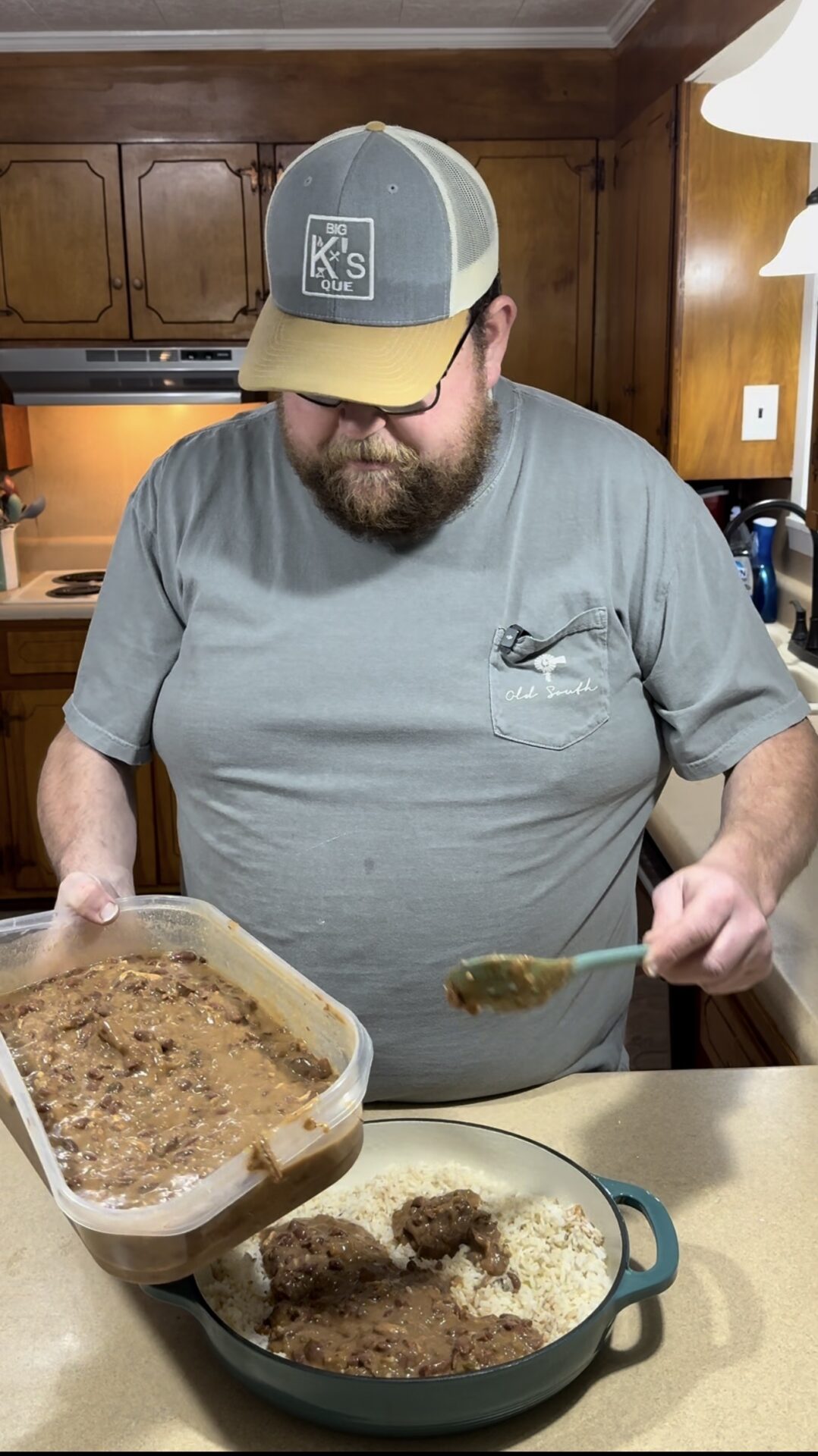 A bearded man in a kitchen scoops brown gravy with meat from a plastic container onto a dish of rice on the counter in front of him. He wears glasses, a gray T-shirt, and a gray and beige cap.