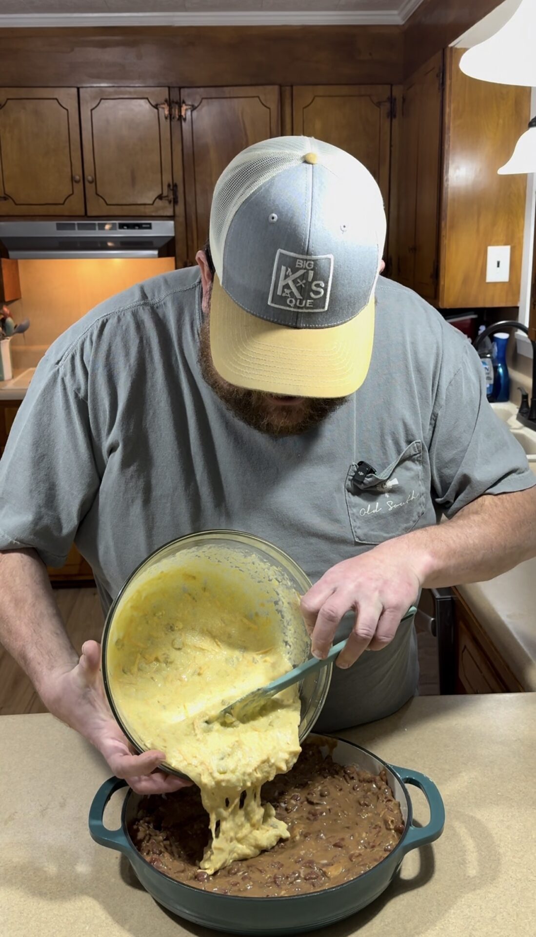A man in a gray shirt and cap pours a bowl of cheesy, yellow casserole mixture over a dish of cooked food in a kitchen with wooden cabinets.
