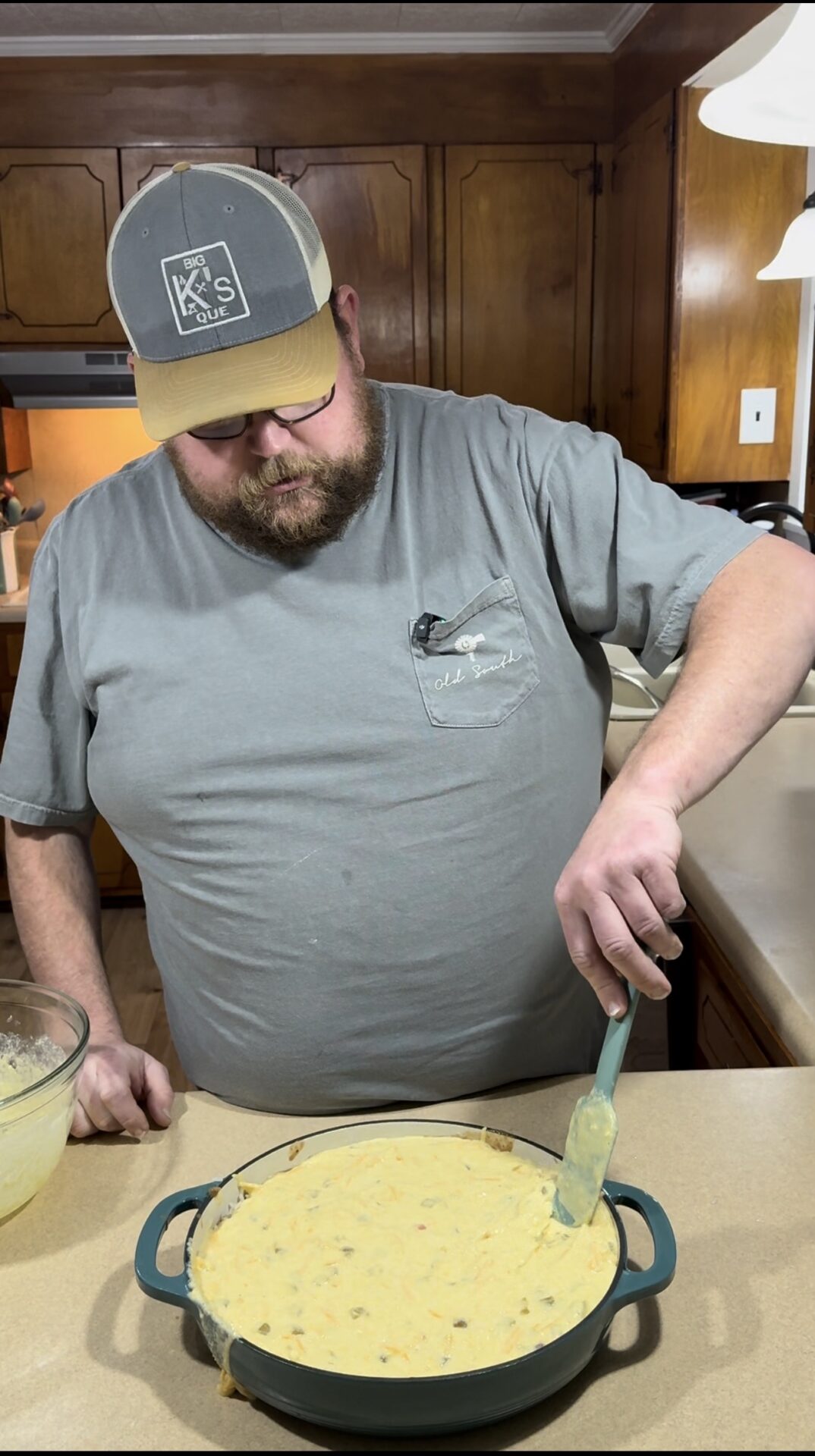 A bearded man in a gray shirt and cap spreads a creamy mixture in a round baking dish with a spatula, standing in a wood-paneled kitchen.