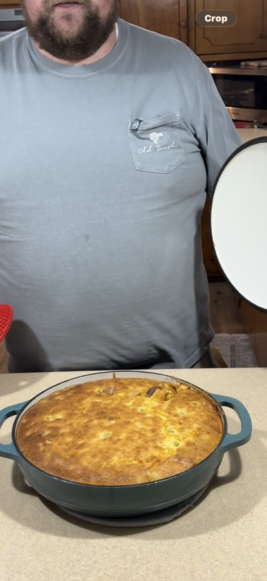 A person in a gray shirt holds the lid of a Dutch oven, revealing a freshly baked, golden-brown casserole inside the pot on a kitchen counter.