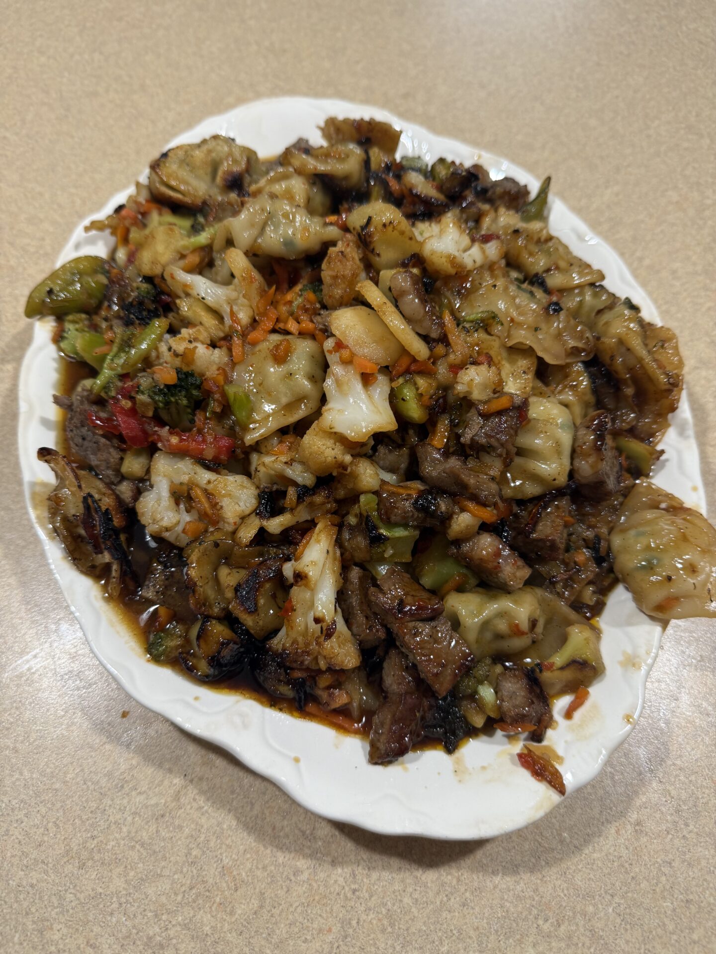 A white oval plate filled with a stir-fried dish containing browned dumplings, chopped vegetables, and slices of beef in a savory sauce, all arranged on a beige countertop.