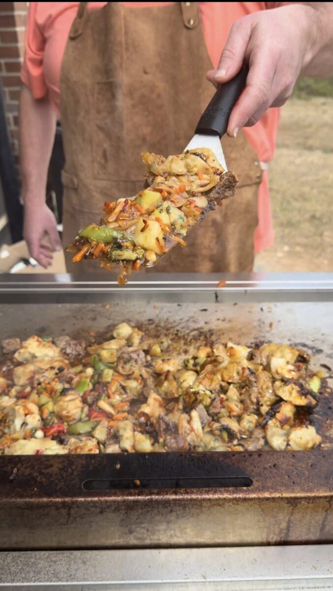 A person wearing a brown apron holds a spatula with grilled vegetables and meat over a hot, flat-top grill loaded with more sizzling food.