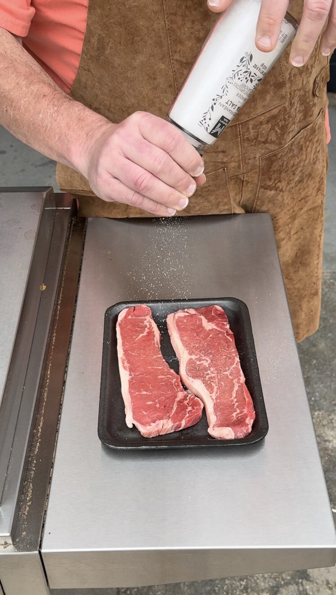 A person wearing a brown apron sprinkles seasoning onto two raw steaks placed on a black tray on a stainless steel surface.