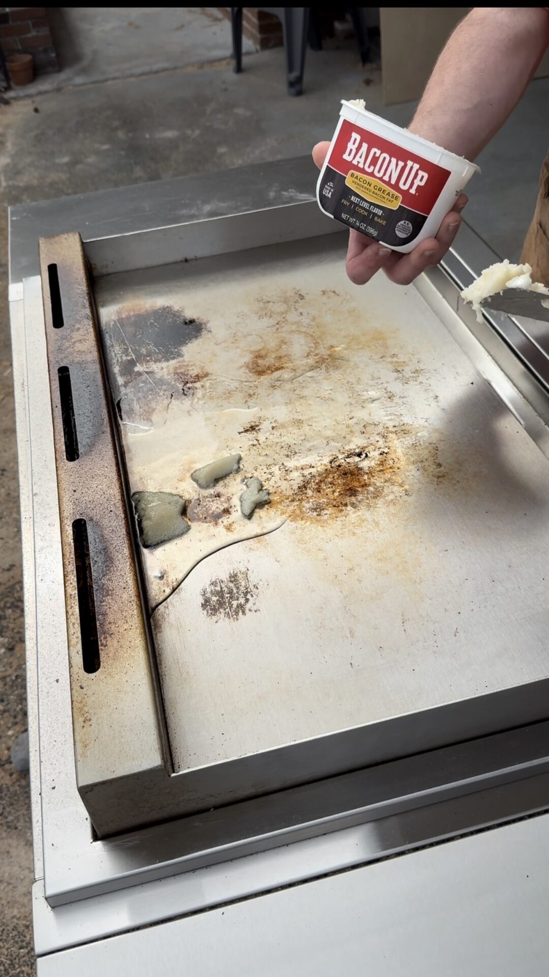 A person spreads Bacon Up bacon grease on a large, slightly dirty outdoor griddle, preparing it for cooking.
