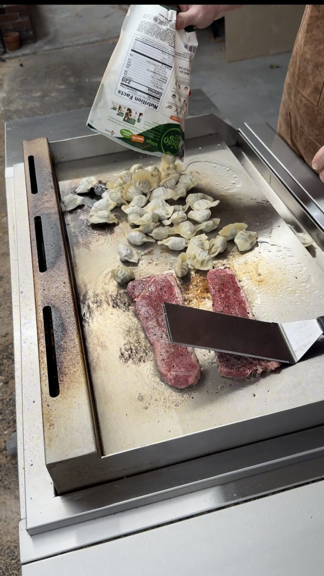 A person cooks two steaks and a pile of frozen dumplings on a flat-top grill. One hand holds a spatula, while the other pours more frozen dumplings from a bag onto the hot surface.