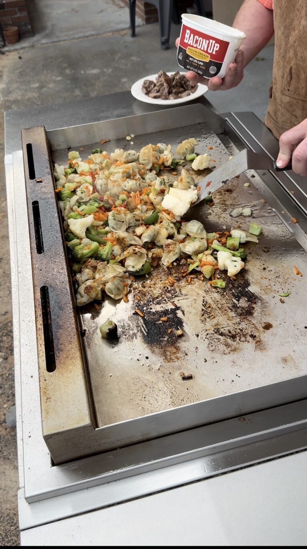 A person is cooking mixed vegetables on a flat-top grill. In one hand, they hold a container labeled Bacon Up and a plate with grilled meat, while the other hand uses a spatula to stir the vegetables.