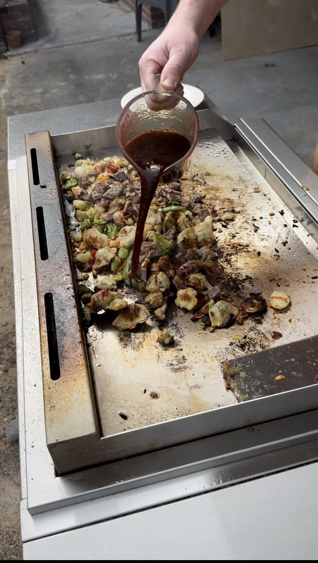 A hand pours dark sauce from a glass measuring cup onto a sizzling mix of vegetables and meat cooking on a flat-top outdoor grill.