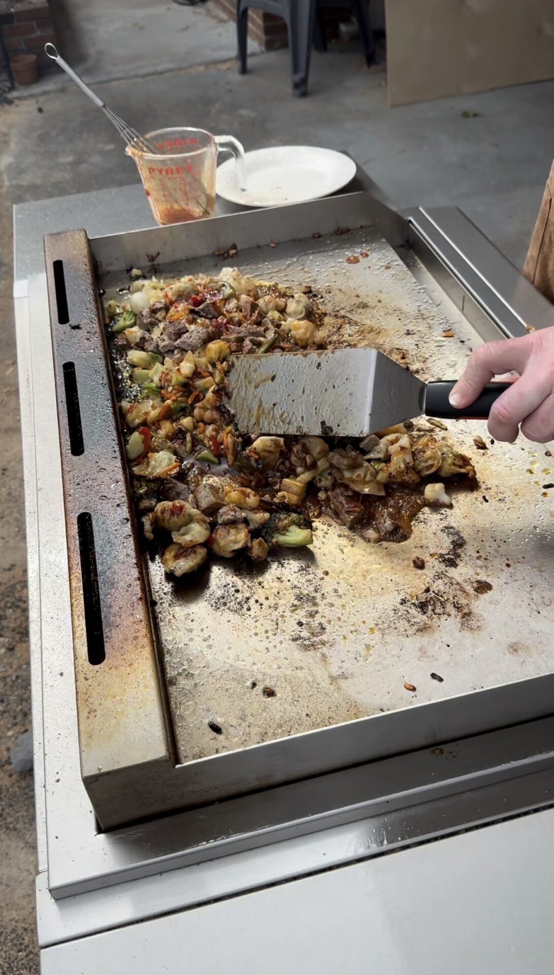 A person uses a spatula to cook and stir a mixture of vegetables and meat on a large outdoor flat-top grill, with a measuring cup and plate in the background.
