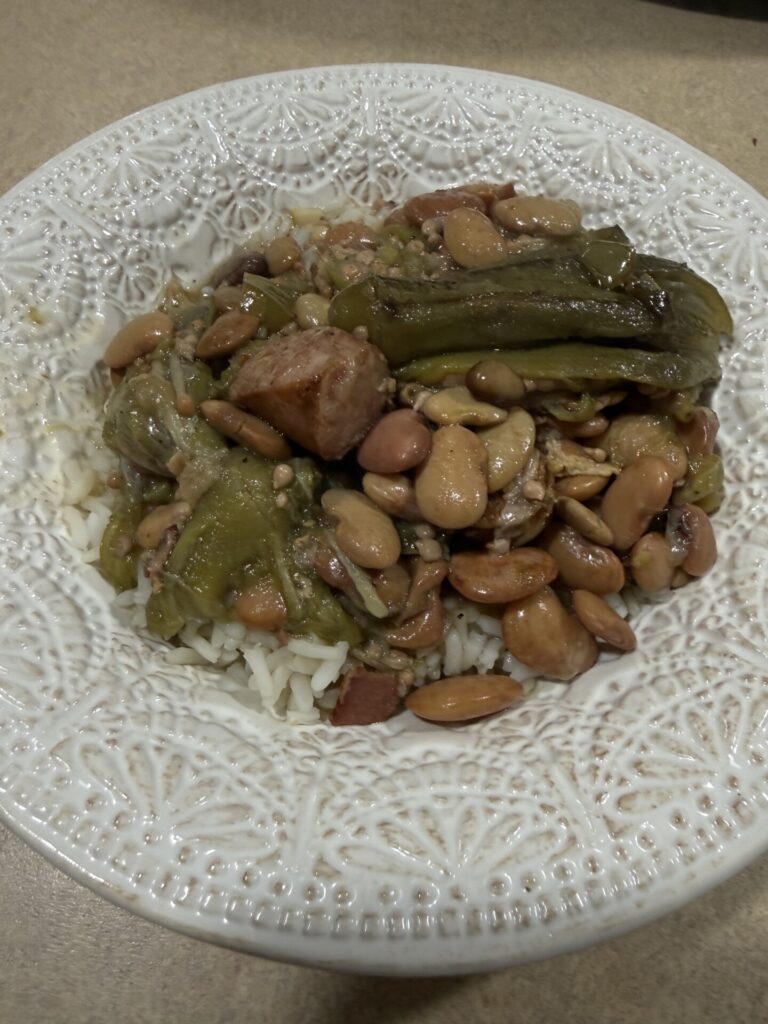 A white patterned bowl filled with white rice topped with beans, sausage pieces, and cooked green vegetables, all mixed together, sitting on a beige countertop.