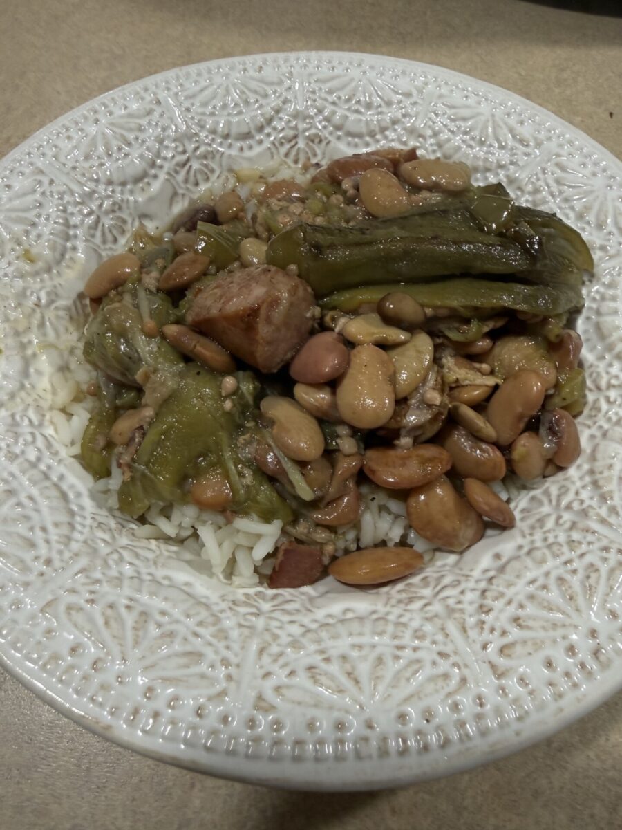 A white patterned bowl filled with white rice topped with beans, sausage pieces, and cooked green vegetables, all mixed together, sitting on a beige countertop.