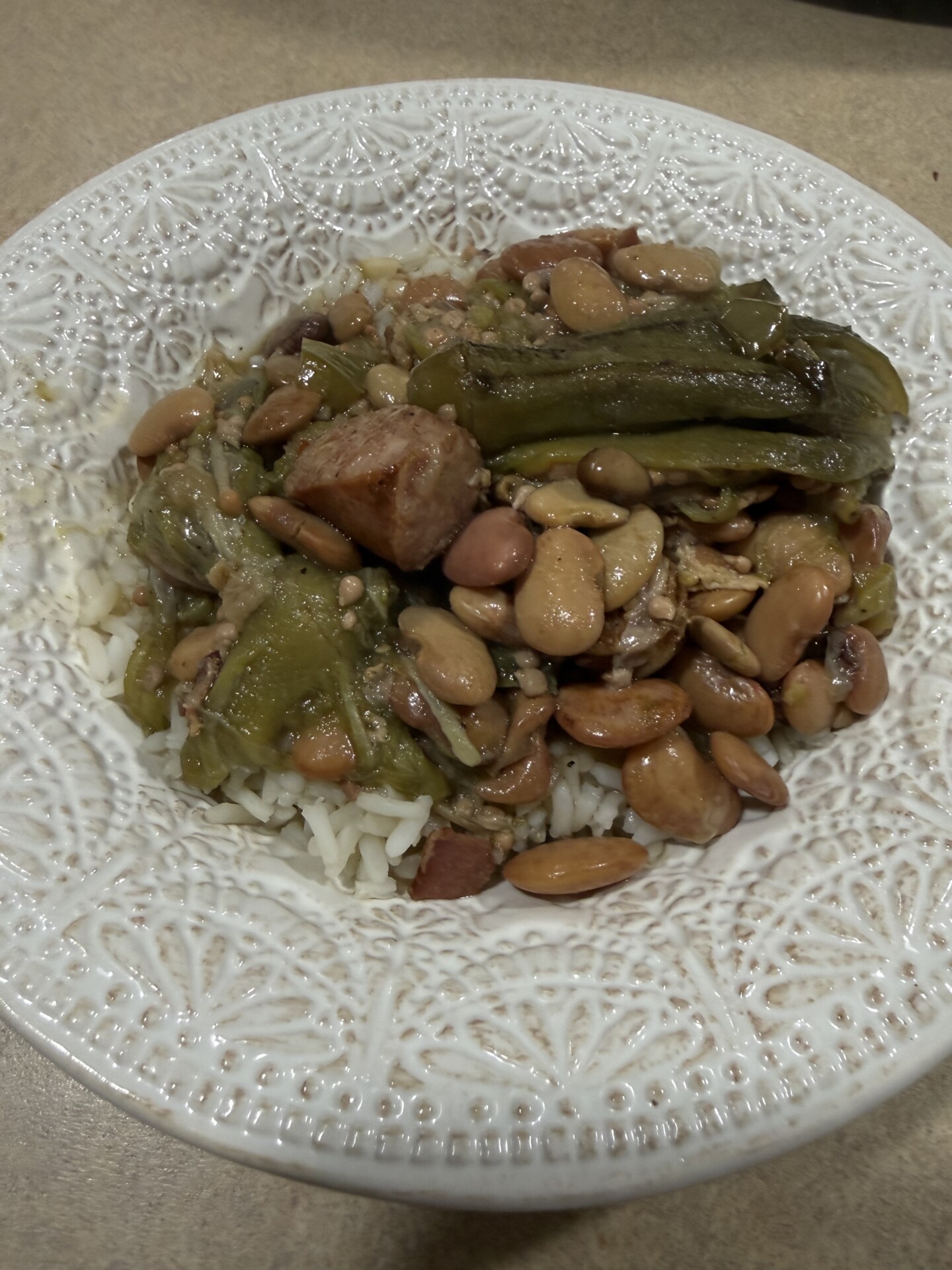 A white patterned bowl filled with white rice topped with beans, sausage pieces, and cooked green vegetables, all mixed together, sitting on a beige countertop.