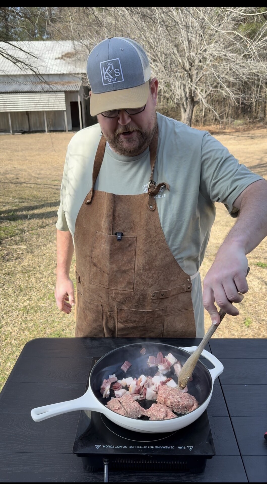 A man wearing a brown apron and tan cap cooks meat in a white pan on an outdoor table, stirring with a wooden spoon. The background shows a grassy yard, trees, and a wooden shed.