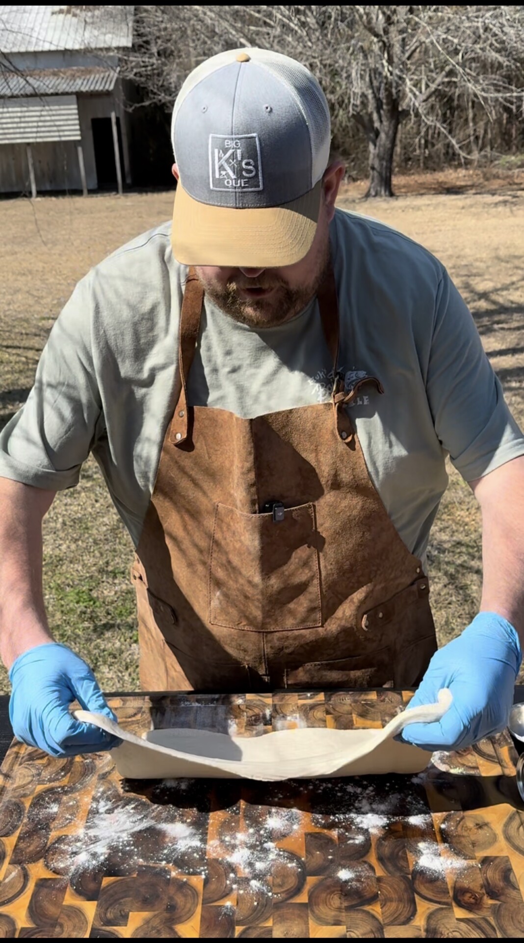 A person wearing a brown apron, blue gloves, and a cap is outdoors, stretching a sheet of dough over a wooden cutting board dusted with flour. Trees and a building are visible in the background.