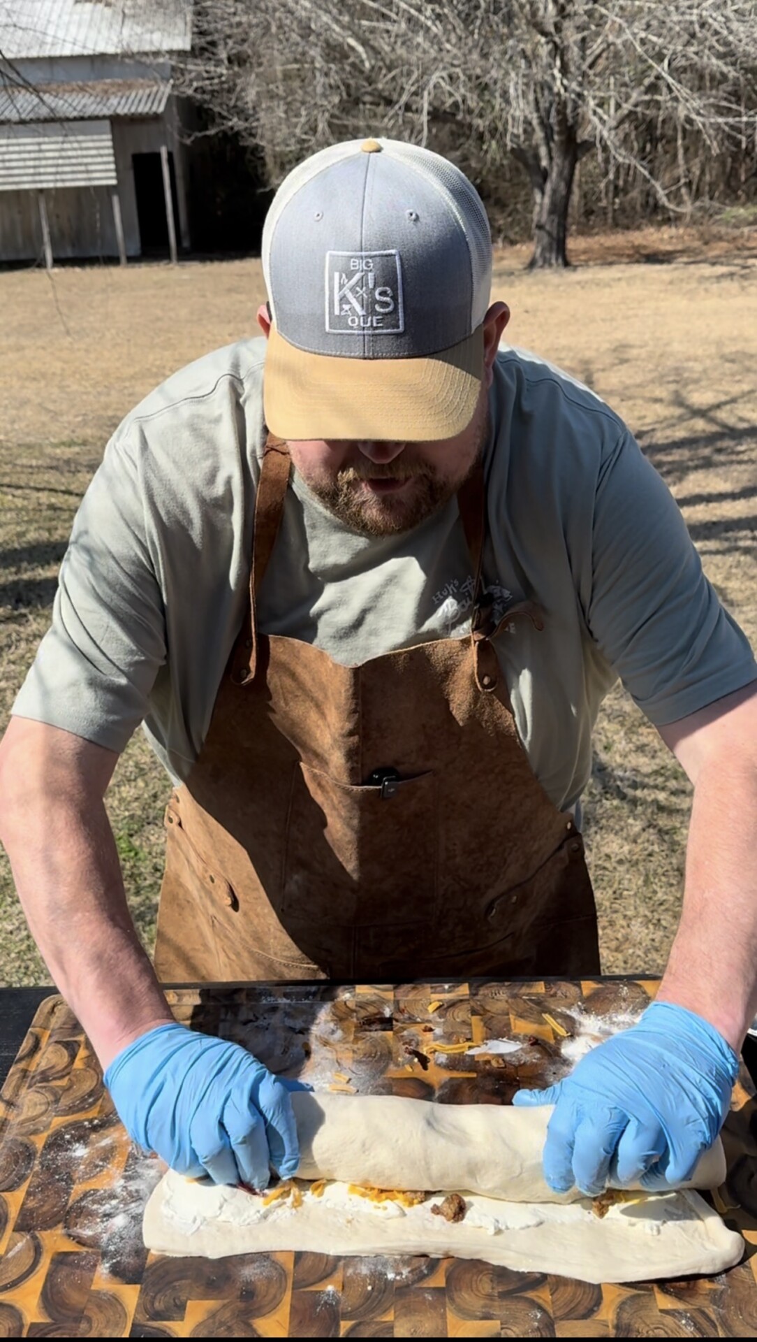 A man wearing a tan apron and blue gloves is rolling dough with filling on a patterned wooden surface outdoors, with dry grass and trees in the background.