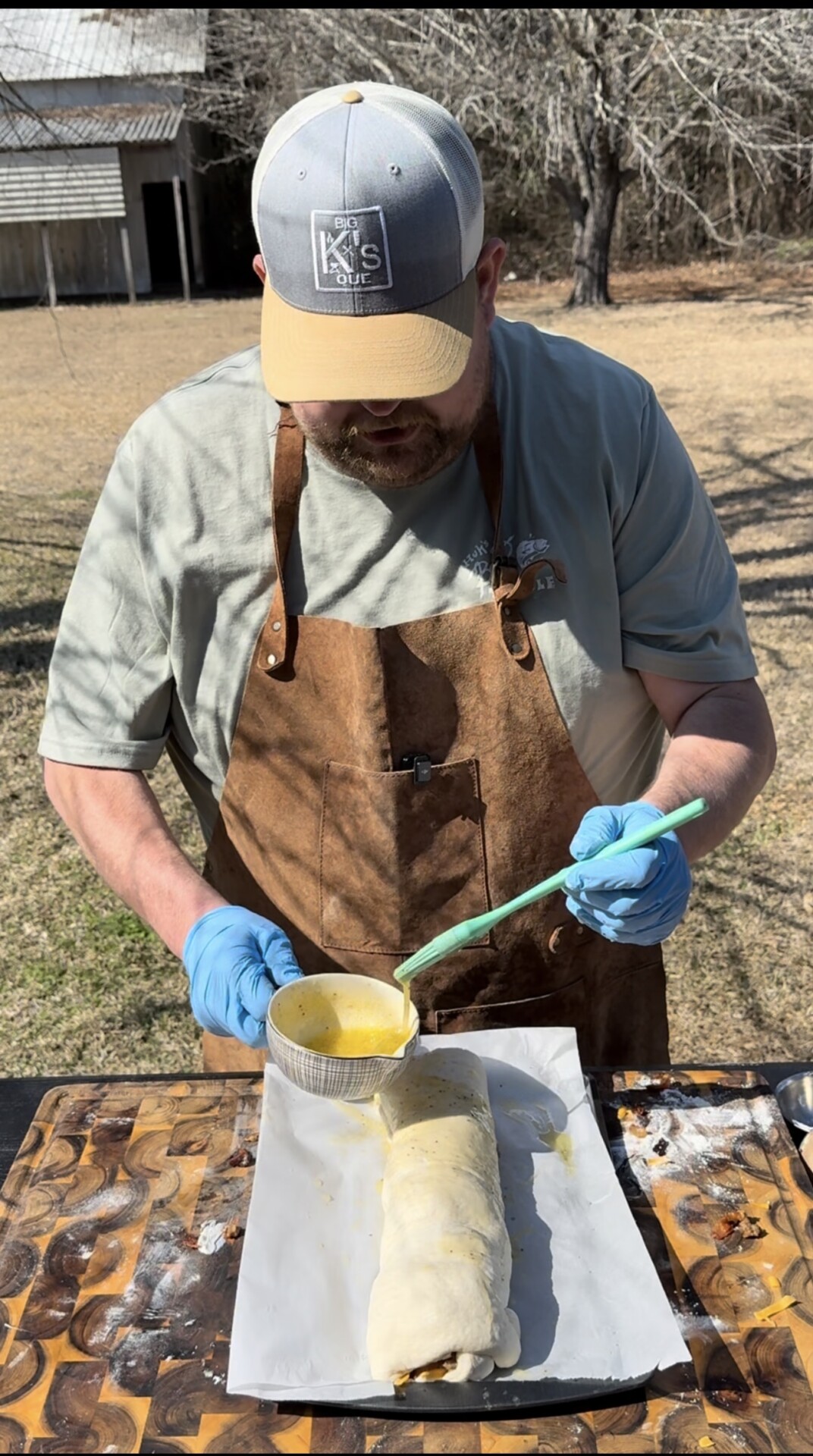 A man wearing a brown apron, blue gloves, and a cap brushes egg wash onto a rolled dough on parchment paper outdoors, with a rustic building and trees in the background.
