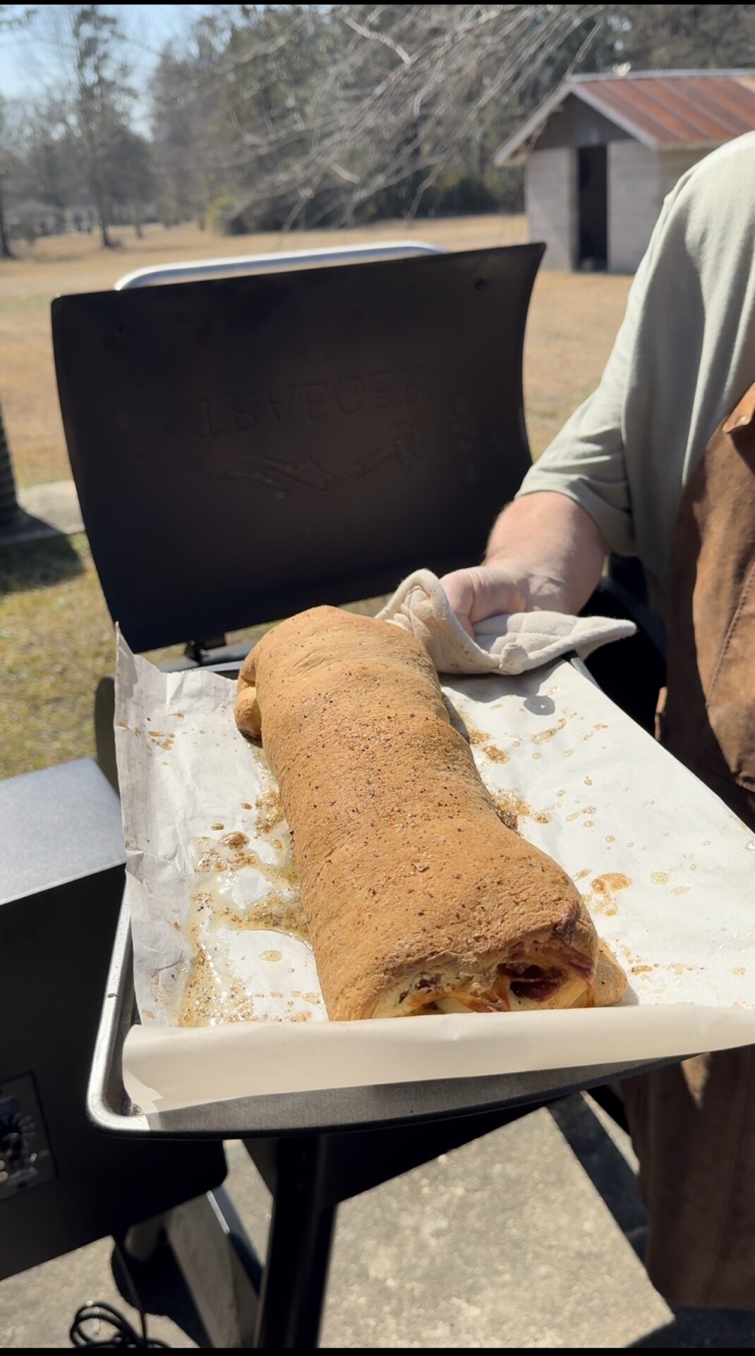 A person wearing an apron holds a baking tray with a large, golden-brown rolled bread fresh from an outdoor oven or grill, set in a sunny backyard.
