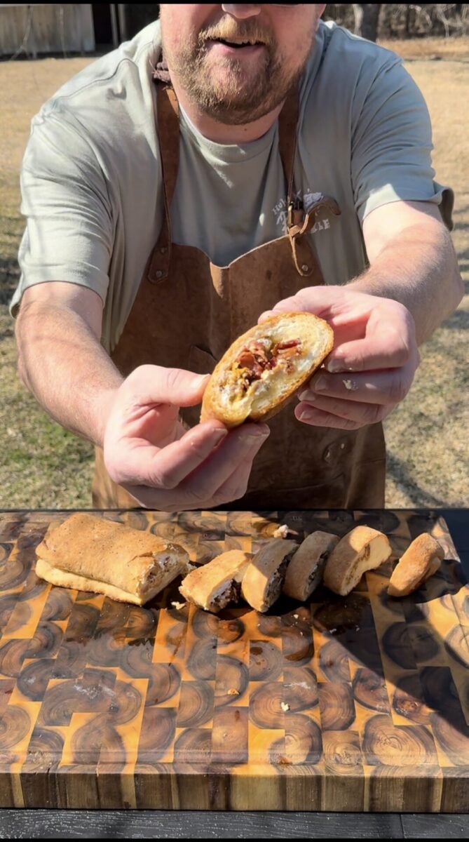 A person wearing a brown apron holds a sliced, stuffed bread roll toward the camera. Several more slices are arranged on a wooden cutting board outdoors. The filling appears cheesy with bits of meat and vegetables.
