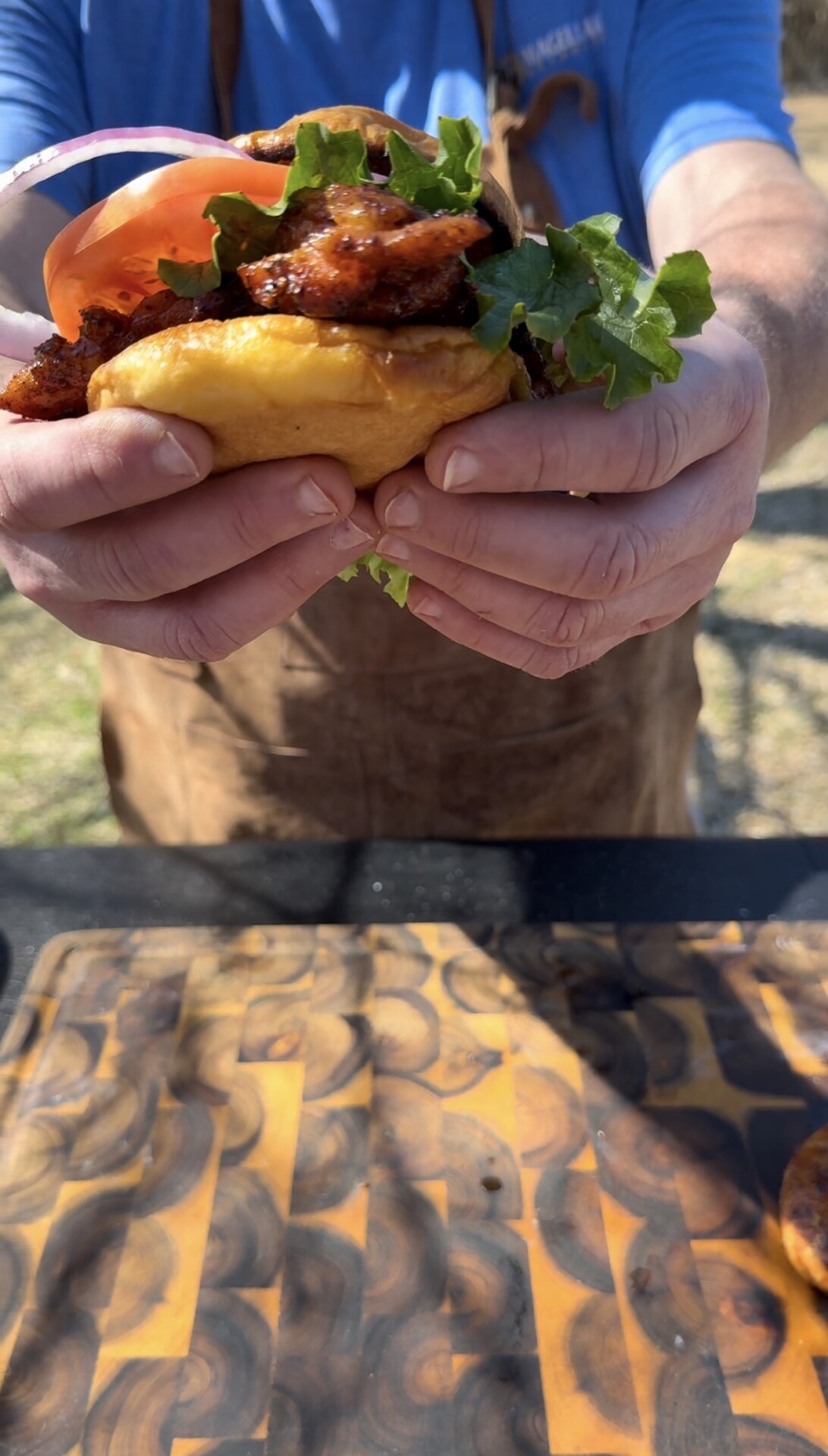 A person wearing a brown apron holds a sandwich filled with fried chicken, lettuce, and tomato. The sandwich is held over a wooden cutting board outdoors in bright sunlight.