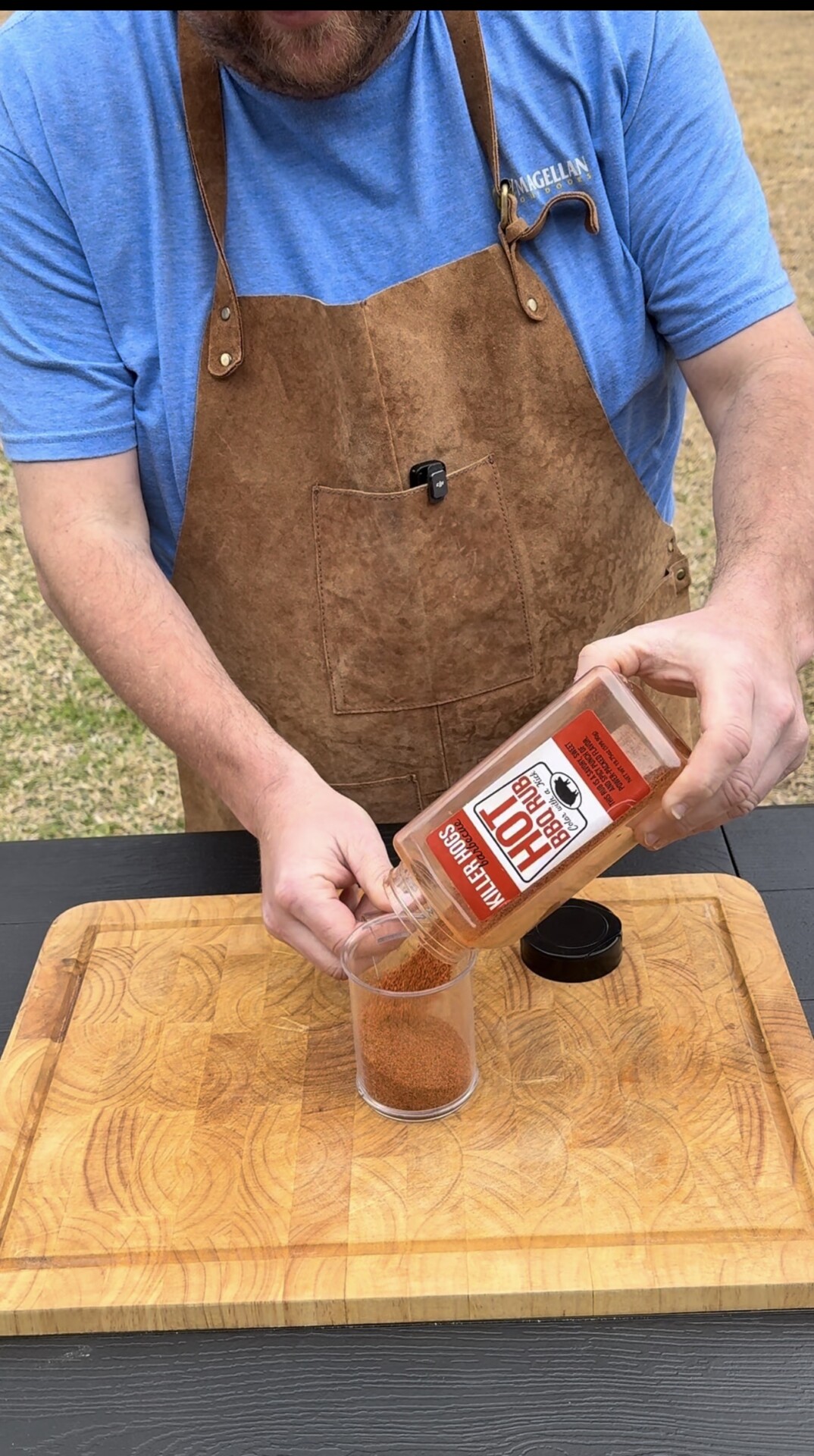 A person in a brown apron pours hot seasoning from a bottle into a clear plastic cup on a wooden cutting board outdoors.