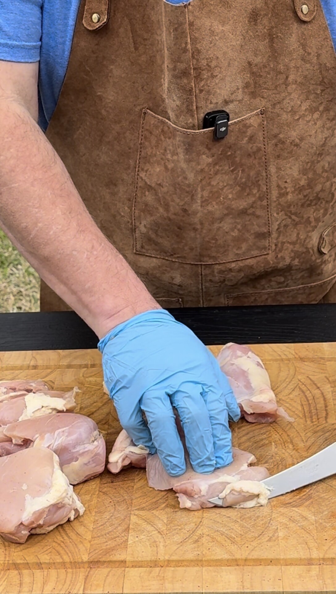 A person wearing a brown apron and blue glove is cutting raw chicken pieces on a wooden cutting board with a knife.