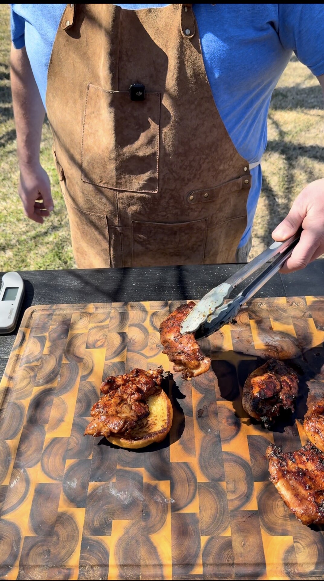 A person wearing a brown apron and blue shirt uses tongs to place grilled meat with sauce onto toasted buns on a patterned wooden cutting board outdoors.
