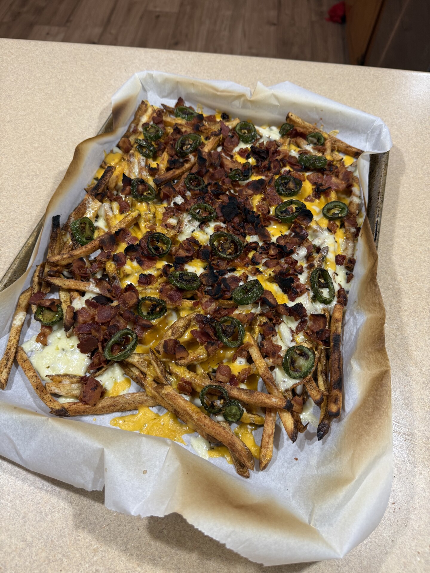 A baking tray lined with parchment paper holds loaded fries topped with melted cheese, bacon bits, and sliced jalapeños, sitting on a kitchen countertop.