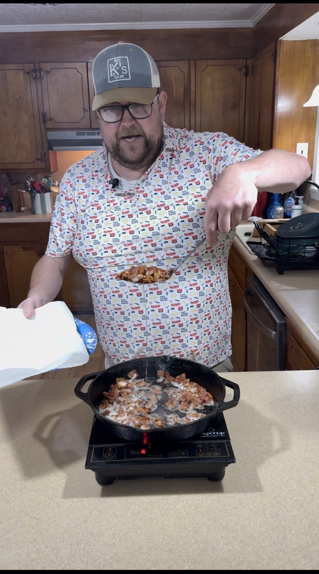 A man in a patterned shirt and cap uses a slotted spoon to lift cooked food from a cast iron skillet on a stove, holding a plate with paper towels in his other hand, in a kitchen with wooden cabinets.