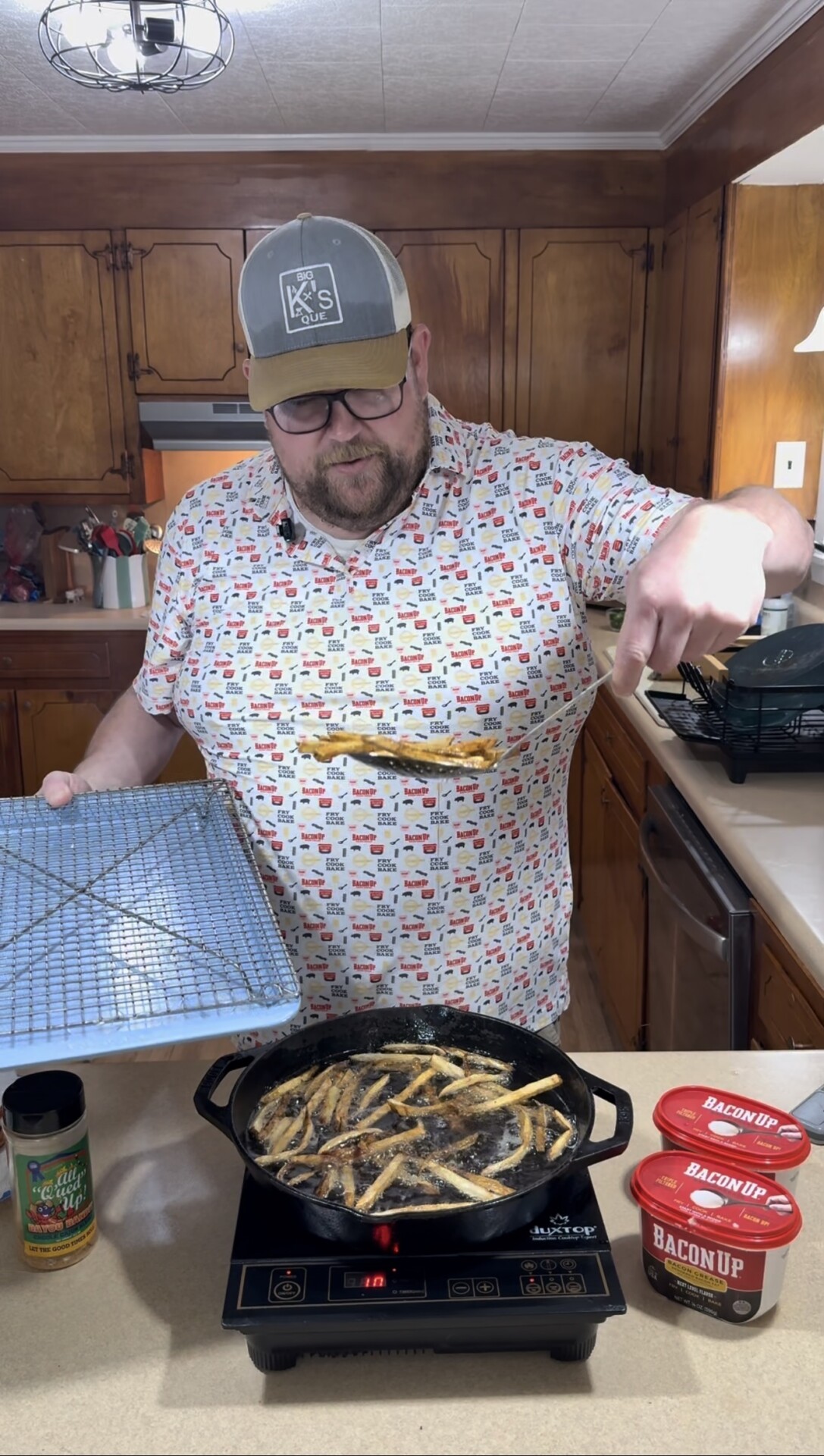 A man in a colorful t-shirt and hat stands in a kitchen, using tongs to cook fries in a cast iron skillet on the stove. He holds a cooling rack in one hand, with seasonings and a bacon grease container nearby.