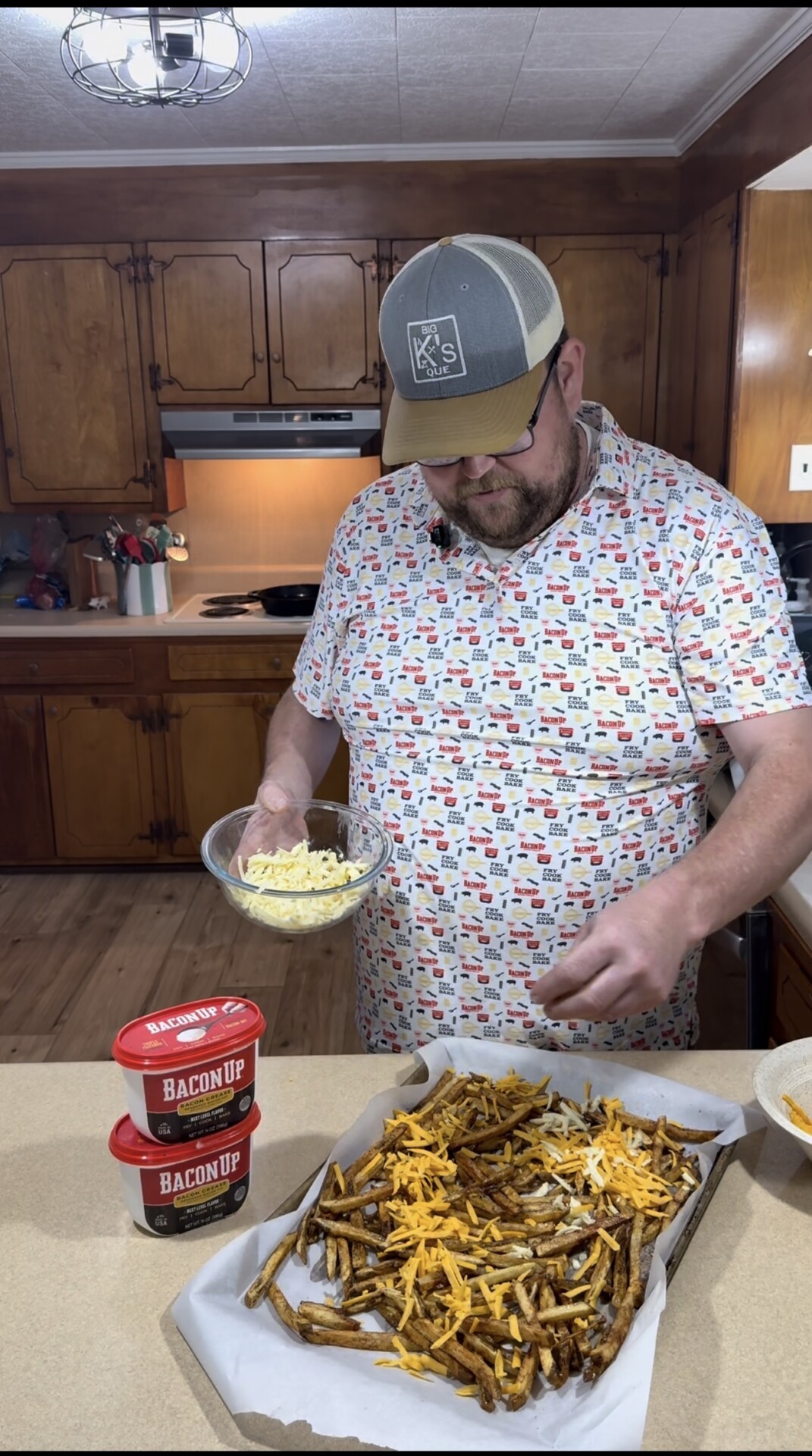 A bearded man in a patterned shirt and cap sprinkles shredded cheese over a tray of fries in a kitchen. Three red containers labeled “Bacon Up” are stacked on the counter next to him.