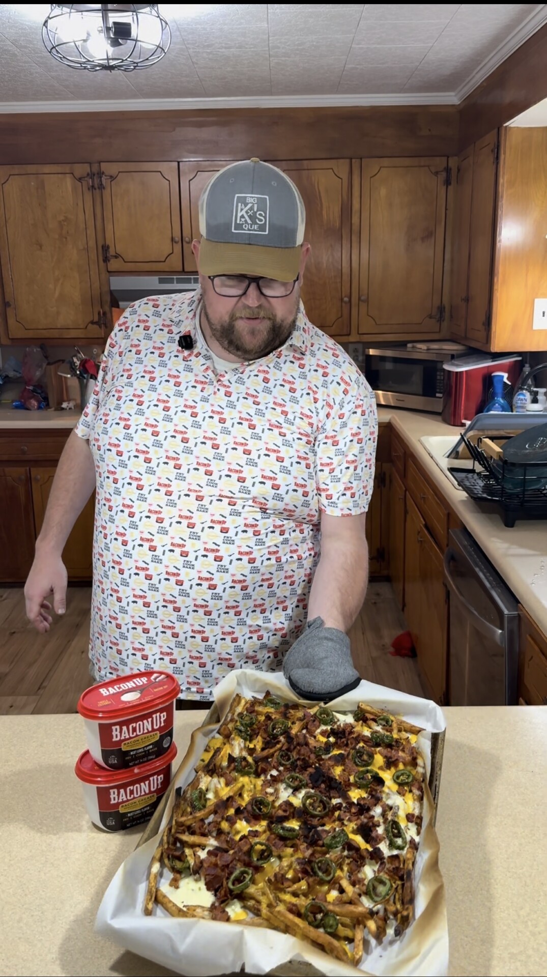 A man wearing a patterned shirt and cap stands in a kitchen, holding a tray of loaded fries topped with cheese, jalapeños, and bacon. Two red containers labeled Bacon Up sit on the counter beside him.