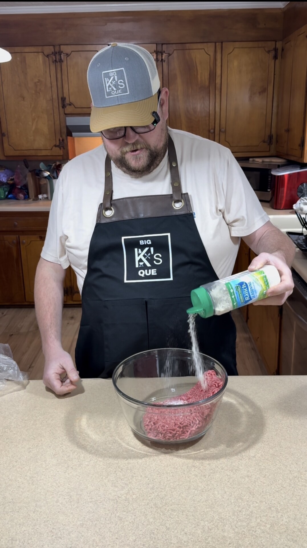 A man wearing a black apron and a baseball cap sprinkles seasoning onto raw ground beef in a glass bowl on a kitchen counter. Wooden cabinets and kitchen appliances are visible in the background.