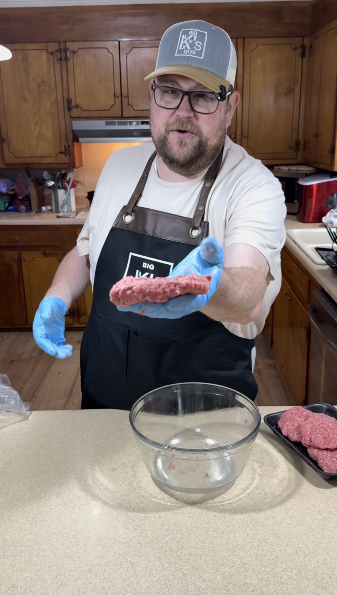 A man wearing a cap, glasses, and gloves holds out a raw hamburger patty in a kitchen, standing behind a counter with a glass bowl and a tray of patties.