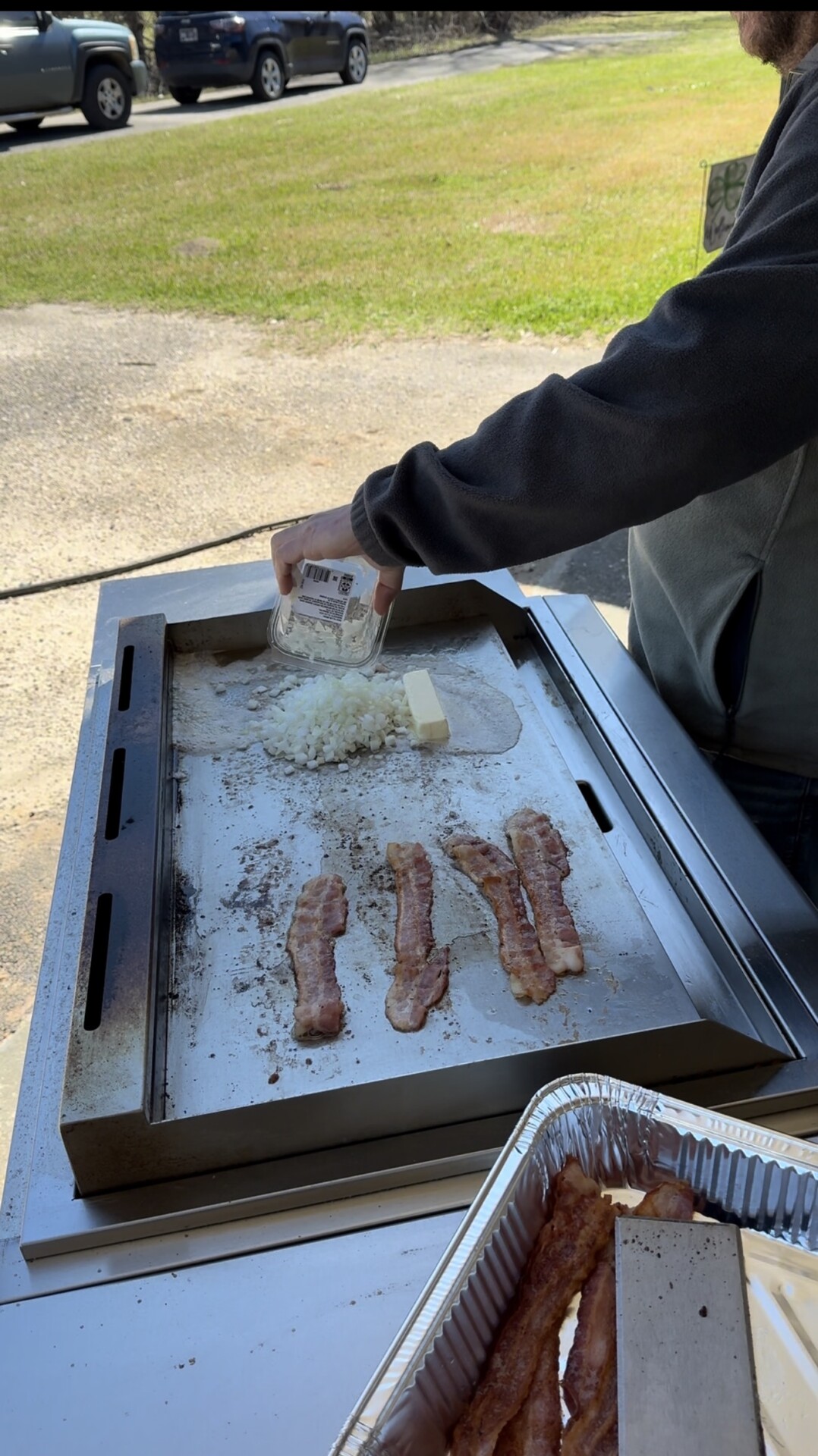 A person pours chopped onions onto a flat-top grill with several strips of bacon and a block of butter, while a tray of cooked bacon sits nearby. The scene is outdoors on a sunny day.