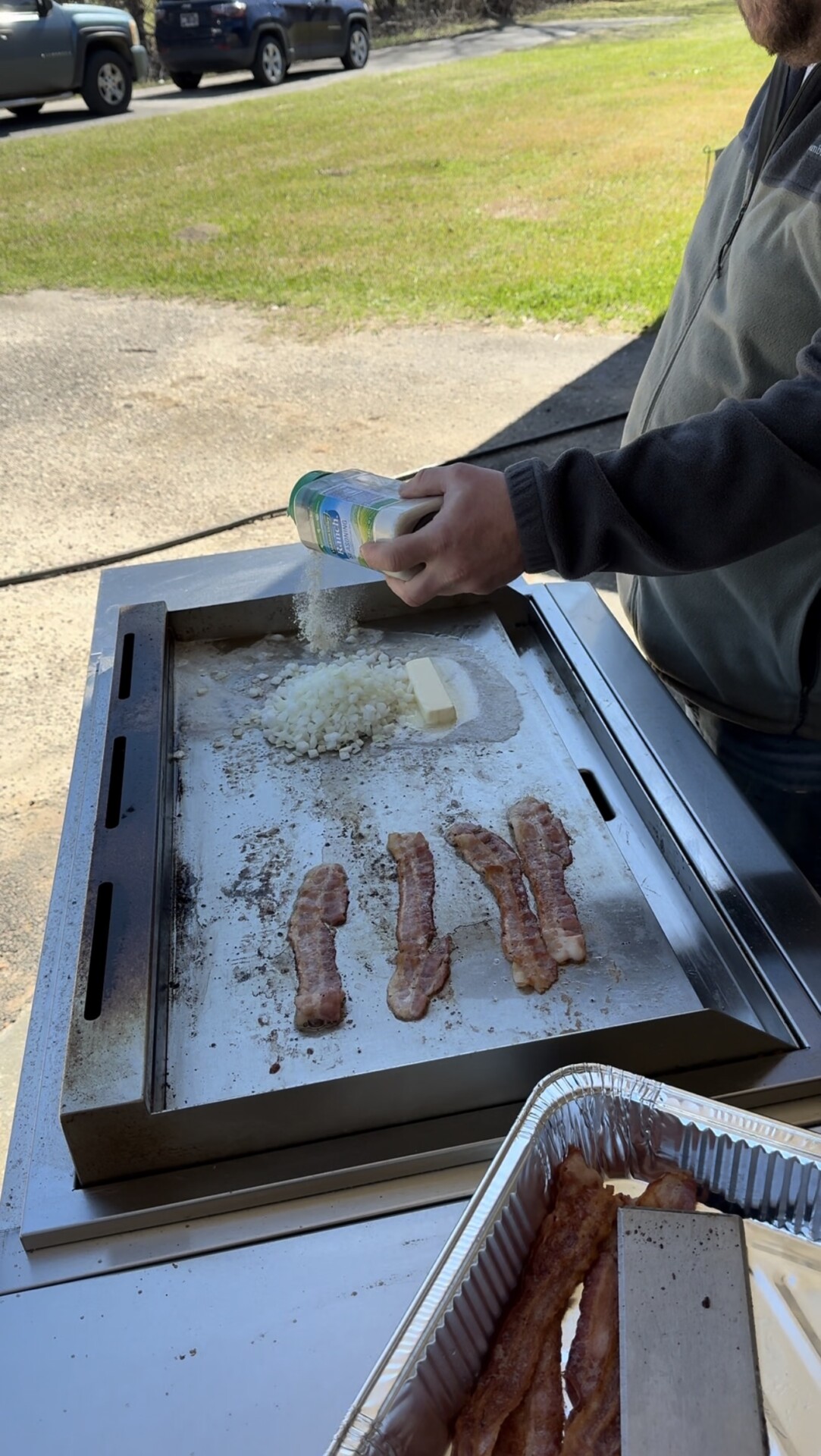 A person sprinkles seasoning onto chopped onions and butter on a griddle, with several strips of bacon cooking nearby. A tray of cooked bacon sits in the foreground, and grass and parked cars are visible in the background.