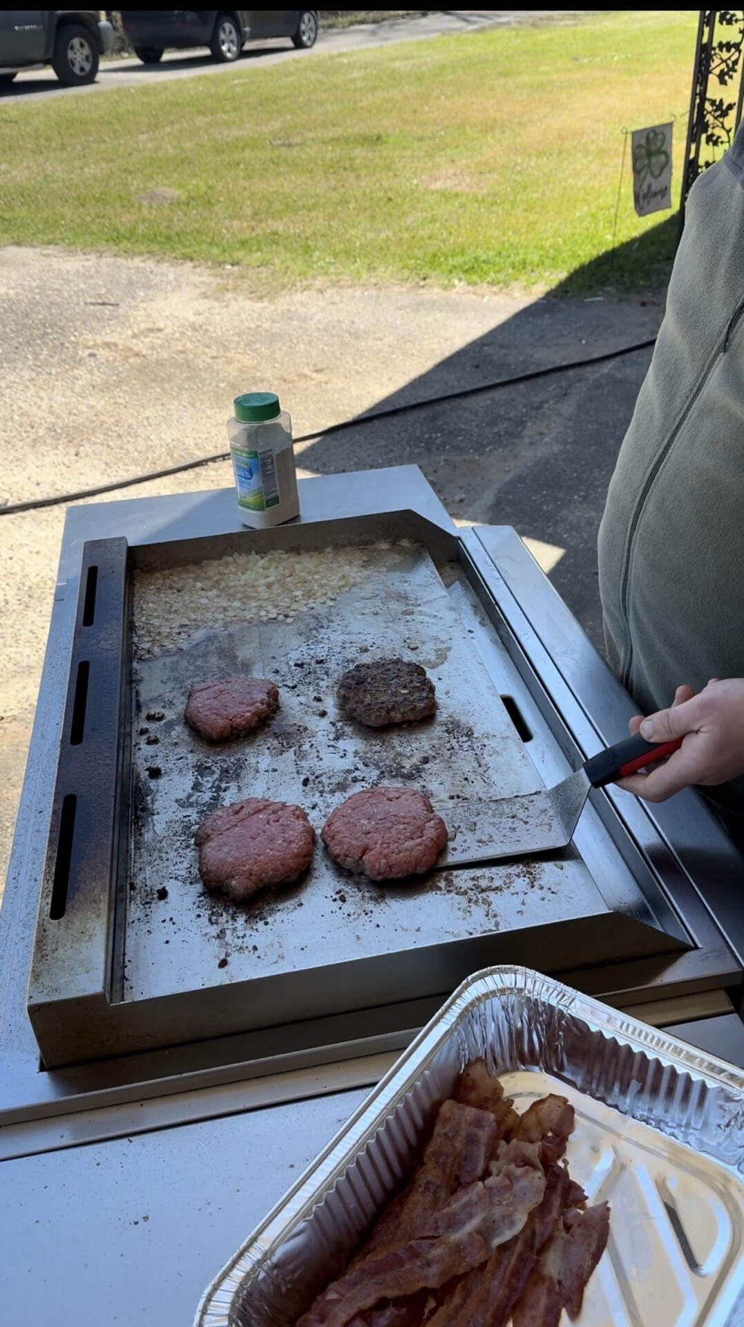 A person flips hamburger patties on a flat-top grill outdoors. Four patties, one cooked, are on the grill, with a tray of uncooked bacon and a jar nearby. Sunlight shines on the scene.