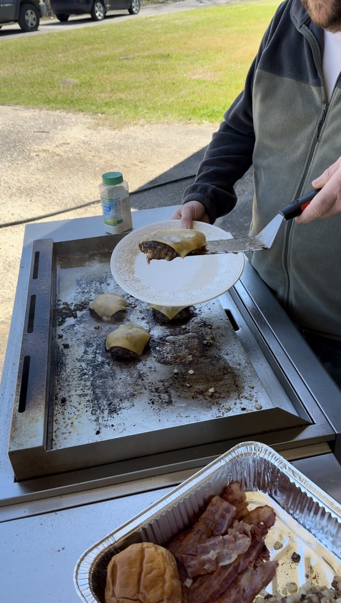A person uses a spatula to place a cheeseburger patty from a griddle onto a white plate. Other patties and cooked bacon are visible nearby, along with a hamburger bun and a seasoning container.
