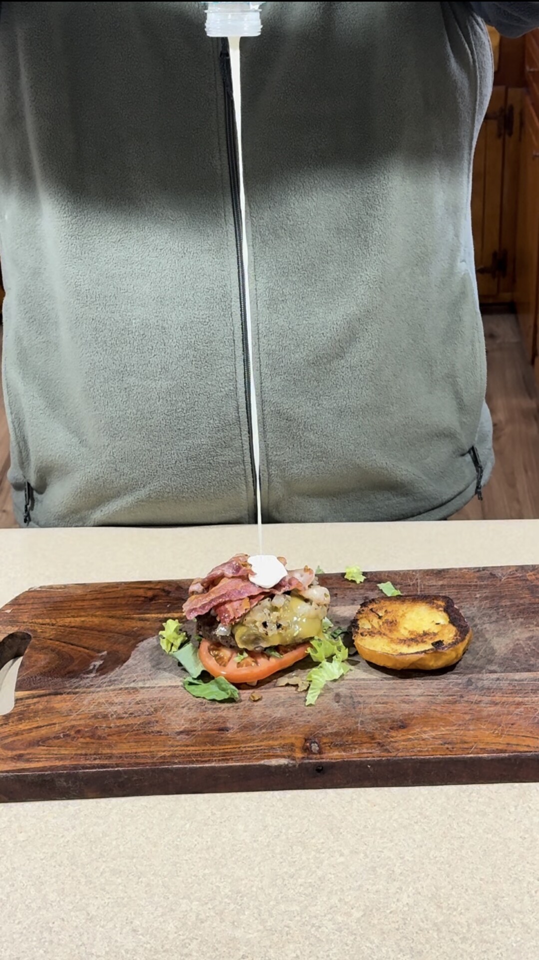 A person in a gray fleece jacket squeezes sauce onto an open burger with lettuce, tomato, and bacon on a wooden cutting board, with the top bun set aside.