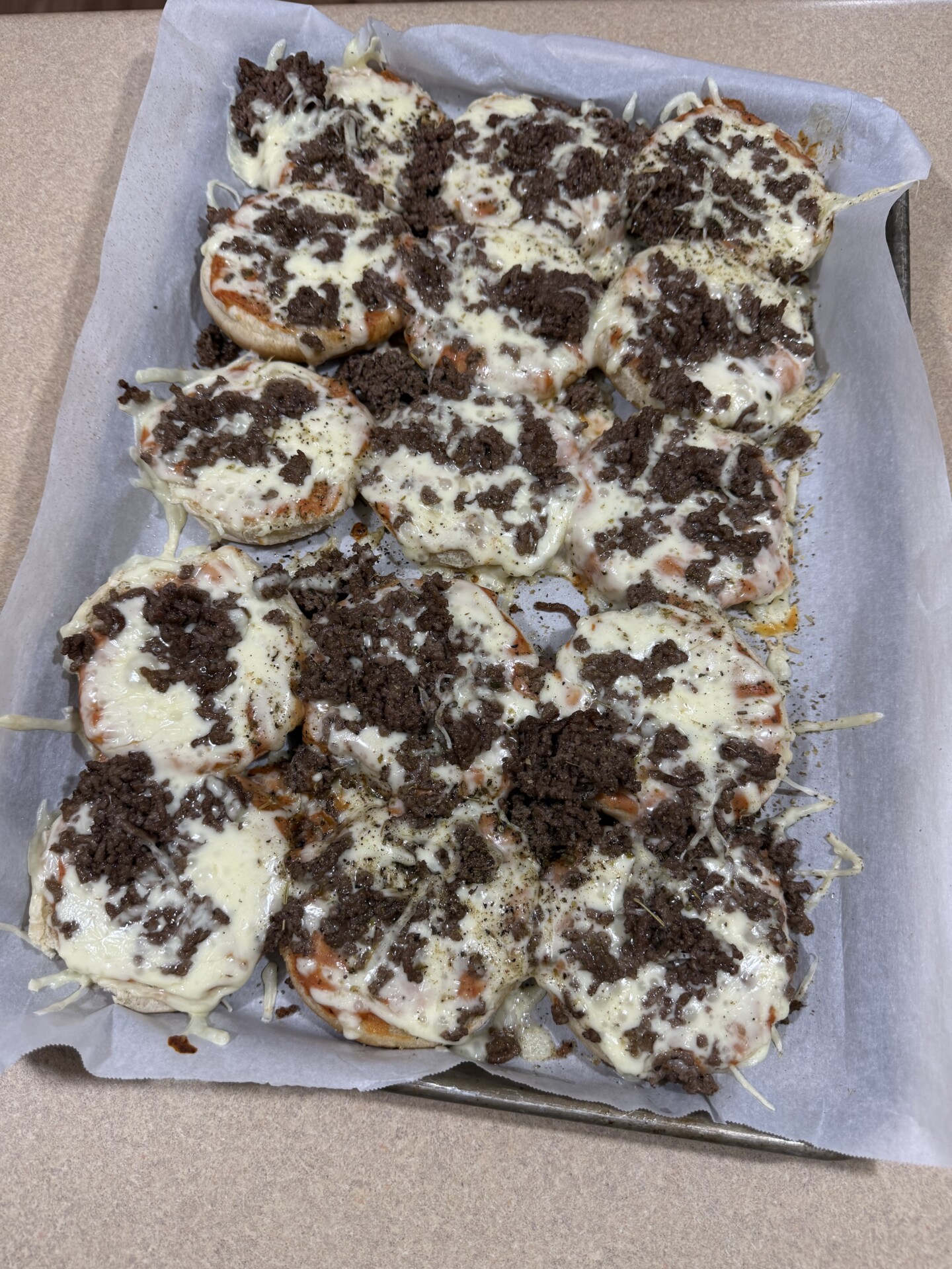 A baking sheet lined with parchment paper holds several mini pizzas topped with melted cheese and crumbled cooked ground beef. The pizzas appear freshly baked and arranged closely together.