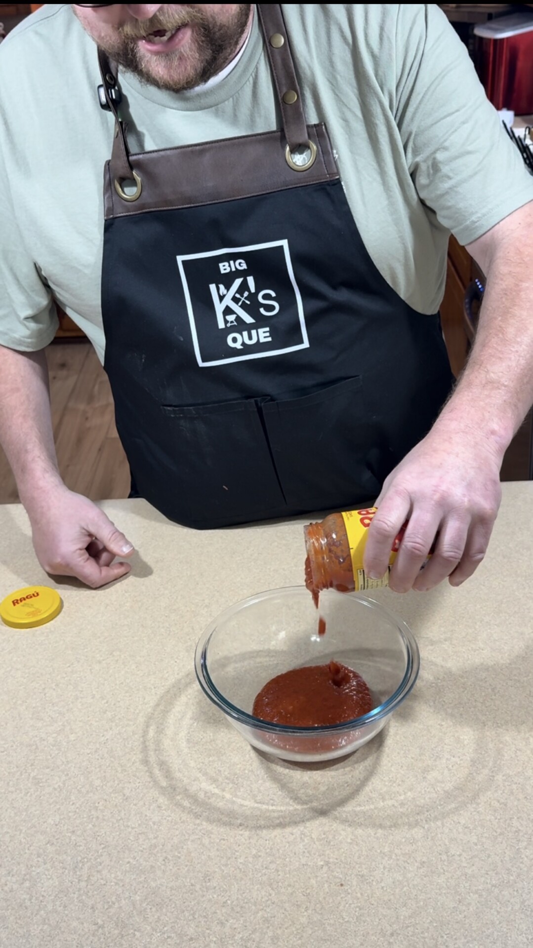 A person wearing an apron labeled BIG KS QUE is pouring barbecue sauce from a jar into a glass bowl on a kitchen counter.
