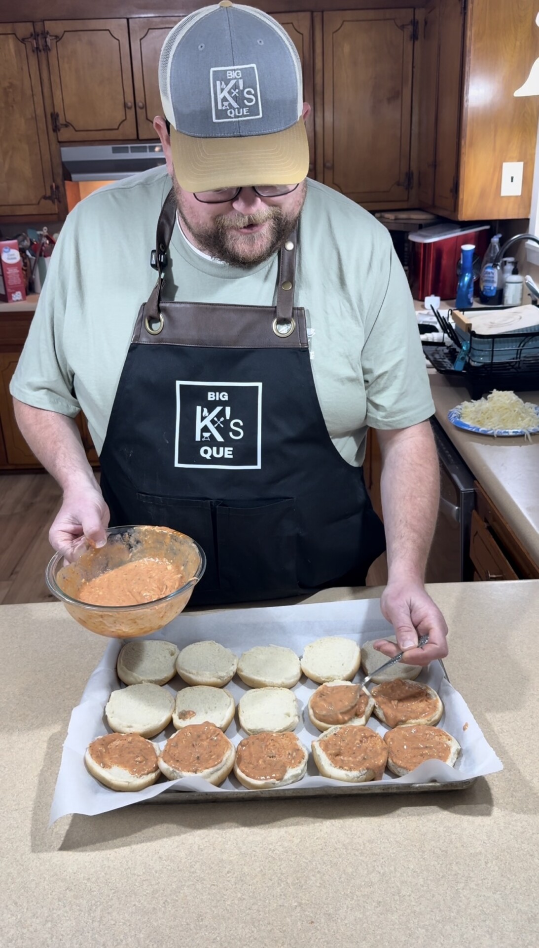 A person wearing a Big Ks Que apron and hat spreads a creamy orange sauce onto open sandwich buns arranged on a baking sheet in a kitchen.