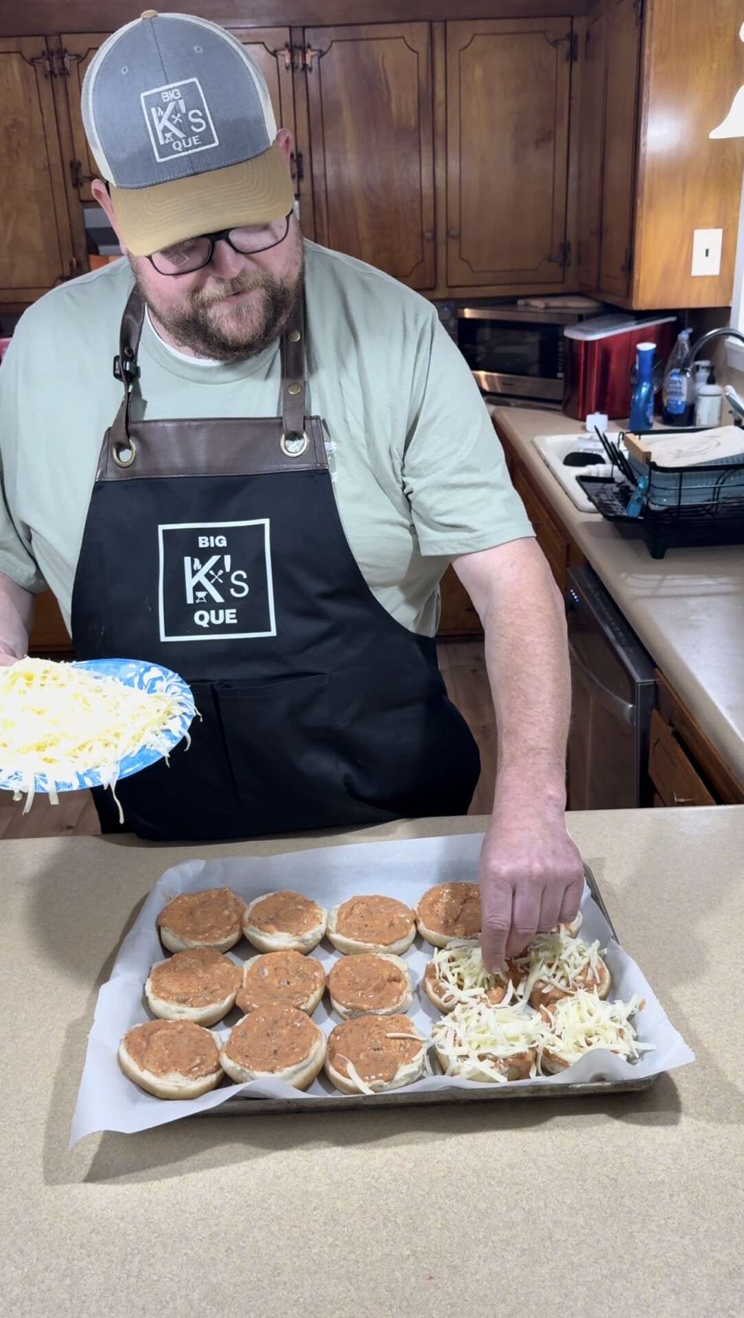 A man wearing a Big Ks Que apron and cap prepares a tray of open-faced sandwiches, sprinkling shredded cheese on top in a home kitchen.