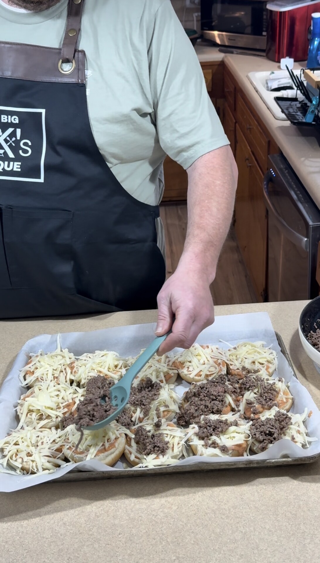 A person wearing a black apron is spooning cooked ground beef onto a tray of slider buns topped with shredded cheese in a kitchen. A skillet with more ground beef sits nearby.