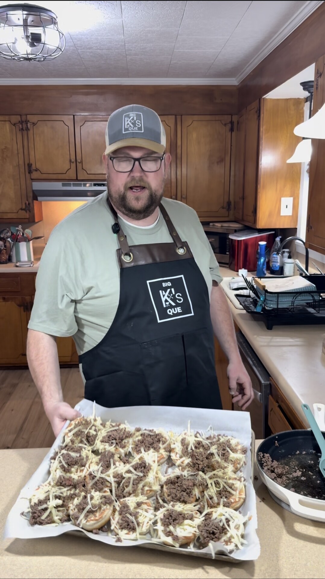 A man in a kitchen wearing a “Big K’s Que” apron and cap holds a tray of open-faced sandwiches topped with shredded cheese and meat, ready to bake. Wooden cabinets and kitchen appliances are visible in the background.