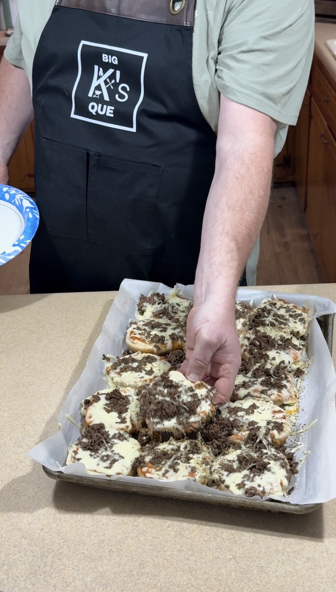 A person wearing a black apron labeled “BIG A’S QUE” lifts a cheesy, ground beef-topped bread slice from a parchment-lined baking tray with several similar pieces on a kitchen counter.