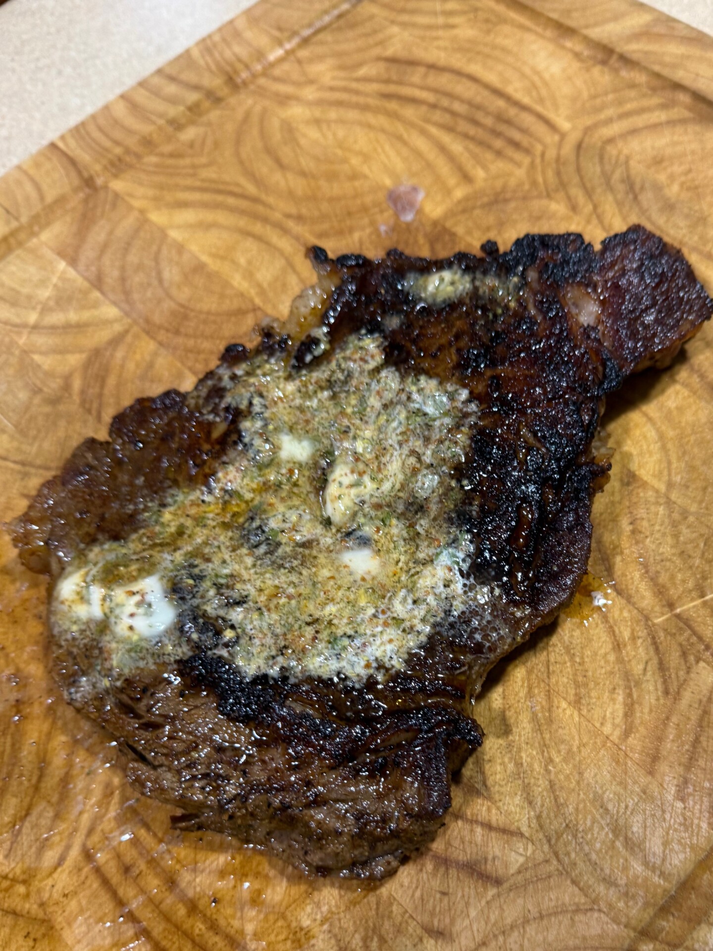 A cooked steak with melted herb butter on top, resting on a wooden cutting board. The steak has a dark seared crust and visible marbling, with juices soaking into the board.